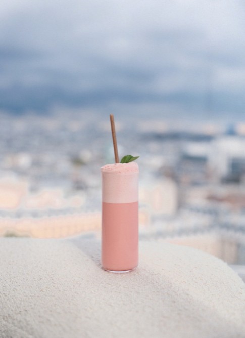 A tall glass of pink frothy drink topped with mint leaves, set against a blurred city skyline background.
