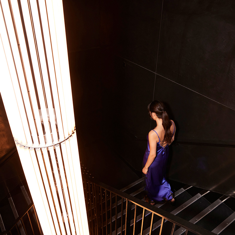 A woman walking down stairs lit only by an illuminated pole at the centre of the staircase
