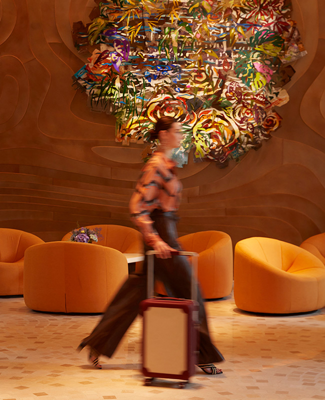 A woman with a suitcase walking past a wall-mounted, resin coloured artwork and orange couches