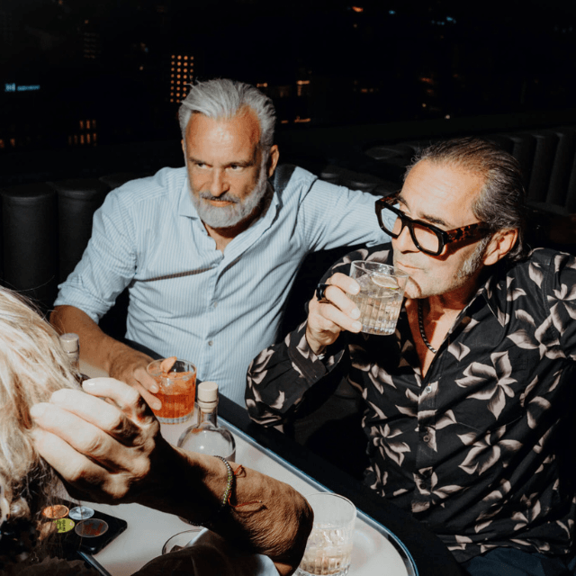 http://Two%20stylish%20gentlemen%20enjoying%20cocktails%20and%20socializing%20at%20a%20sophisticated,%20dimly-lit%20rooftop%20bar%20at%20night.
