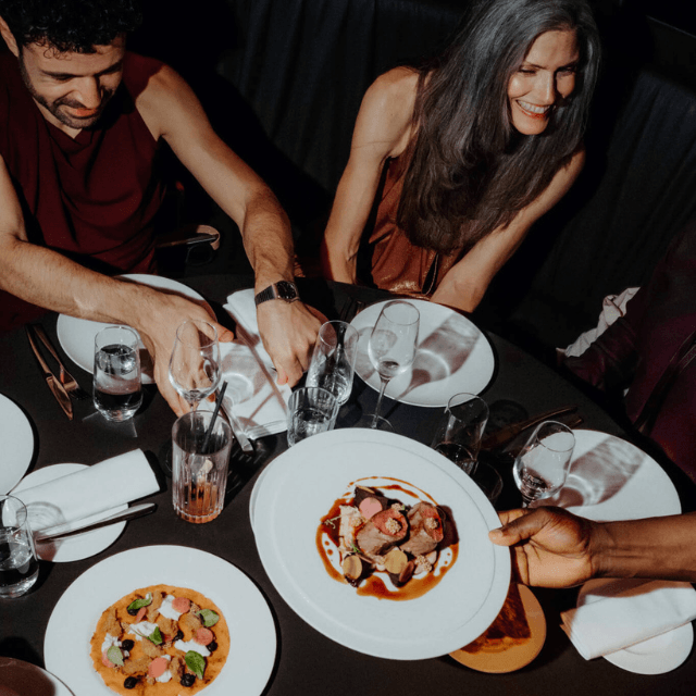 http://Top-down%20view%20of%20an%20elegantly%20plated%20meat%20dish%20with%20rich%20jus%20and%20roasted%20vegetables%20being%20served%20to%20guests%20on%20a%20dark%20wooden%20table.