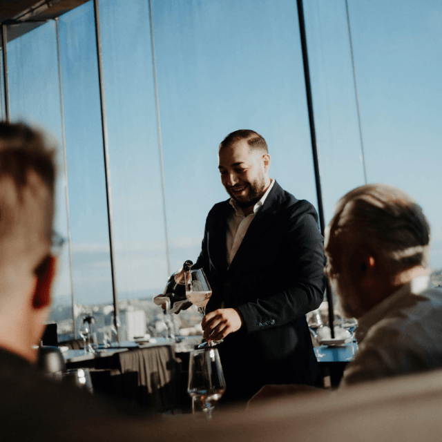 http://A%20professional%20sommelier%20in%20a%20suit%20smiling%20while%20pouring%20champagne%20from%20a%20wrapped%20bottle%20for%20guests%20at%20an%20upscale%20restaurant%20with%20floor-to-ceiling%20panoramic%20windows.