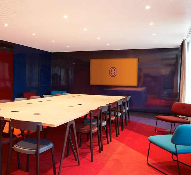 A long meeting table and chairs is set up in front of a screen, hung on a blue wall.
