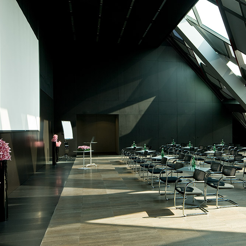A large, high ceilinged meeting room. Tables and chairs in a classroom set up face large screen hanging on the wall.