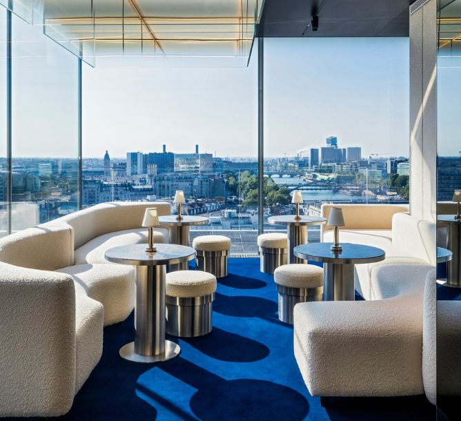 Modern lounge with plush white sofas, round tables, and cityscape view through large windows. Vibrant blue carpet adds a serene touch.