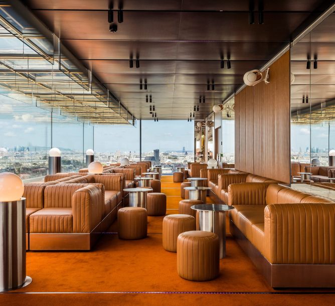 Luxurious rooftop lounge with tan leather seating and round stools on orange carpet. Glass walls offer panoramic city views, creating a modern, elegant atmosphere.