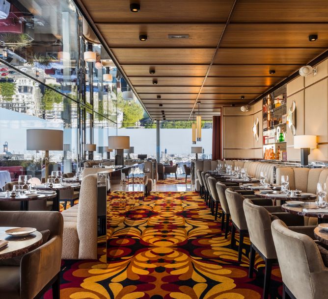 Elegant restaurant interior with floor-to-ceiling windows, mirrored ceiling, colorful floral carpet, and neatly set tables, creating a luxurious ambiance.