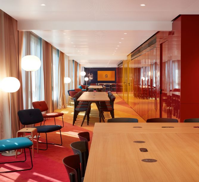 Modern conference room with long wooden tables, colorful chairs, and red walls. Soft lighting from large globe lamps creates a warm, inviting atmosphere.