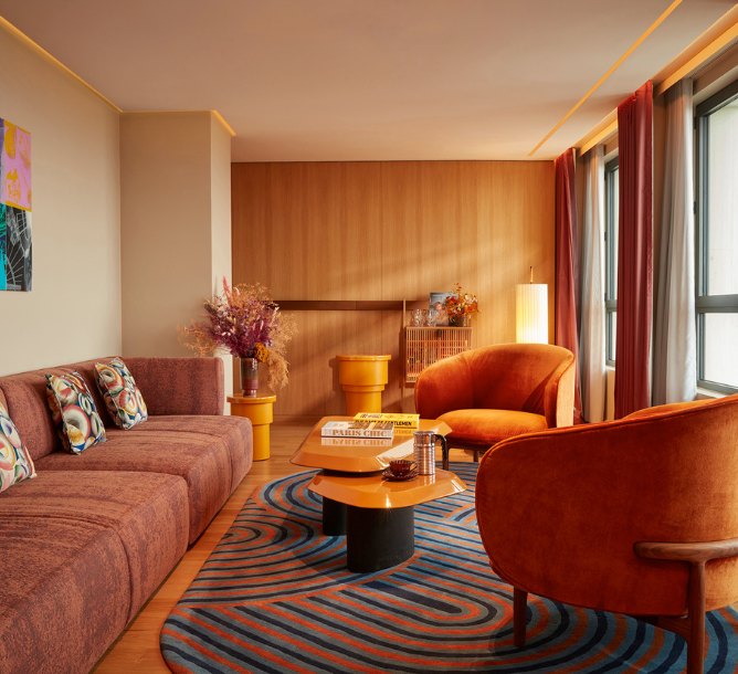 Warm, inviting living room with orange armchairs, patterned brown couch, vibrant cushions, geometric rug, and soft lighting, creating a cozy ambiance.