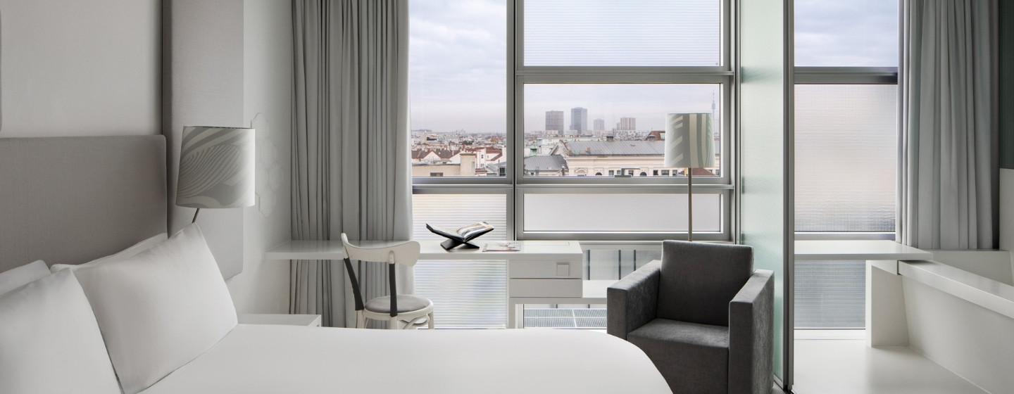 Modern hotel room with a minimalist design featuring a large bed, sleek chair, and desk. Large windows offer a city view, creating an airy, tranquil ambiance.