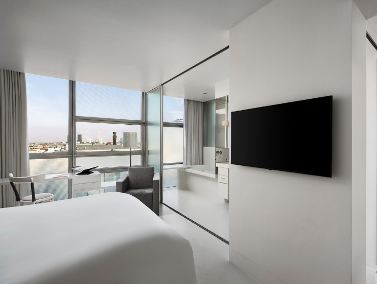 Minimalist hotel room with a large bed, flat-screen TV, glass-walled bathroom, modern desk with a chair, and city view through tall windows.
