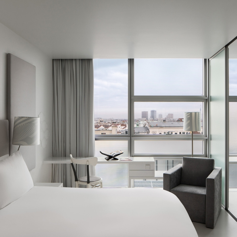 Modern hotel room with a minimalist design featuring a large bed, sleek chair, and desk. Large windows offer a city view, creating an airy, tranquil ambiance.