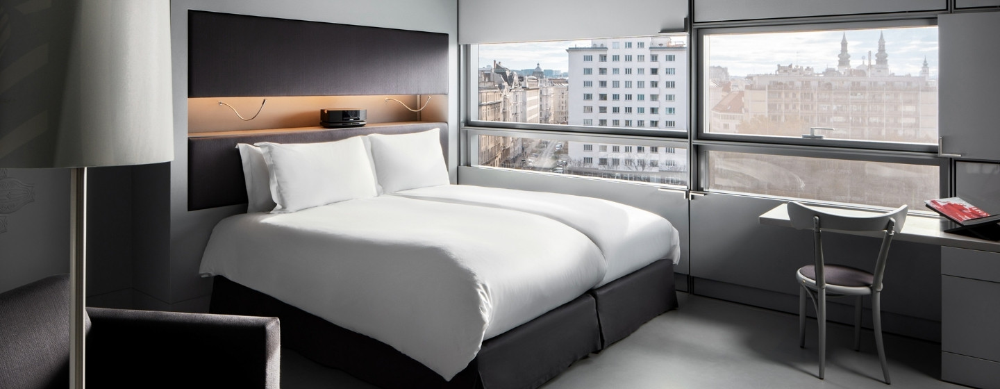 Modern hotel room with a plush double bed, sleek gray walls, and large windows showcasing a cityscape. A desk with a chair adds a functional touch.