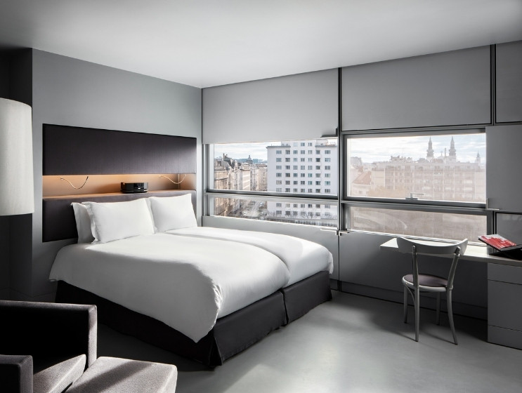 Modern hotel room with a large bed, crisp white linens, gray walls, and a city view through wide windows. Cozy armchair and sleek desk complete the scene.