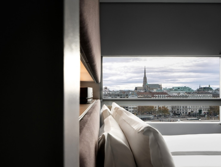 Modern hotel room with crisp white pillows on a bed, framed by a large window. View showcases a city skyline with a prominent church steeple.