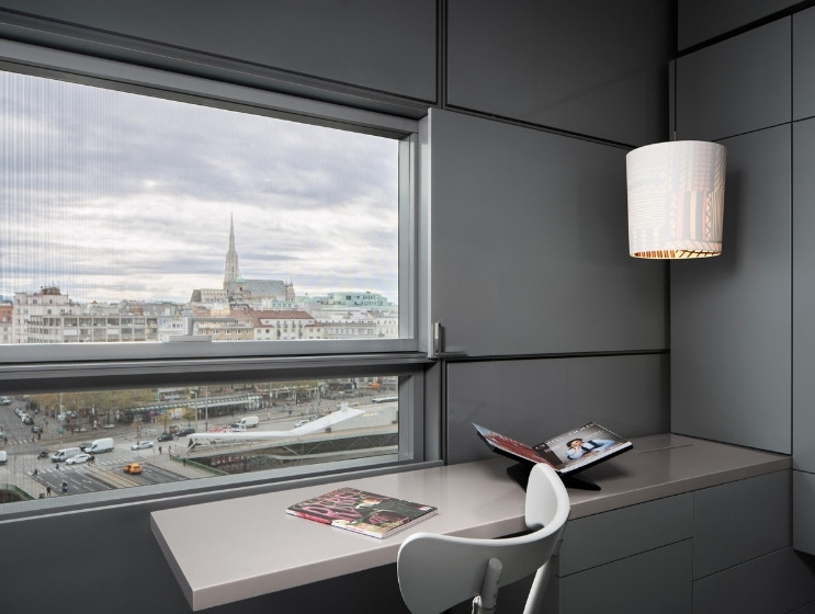 A sleek modern office with gray paneled walls and a large window revealing a city view with a church spire. A white chair faces a desk with magazines.