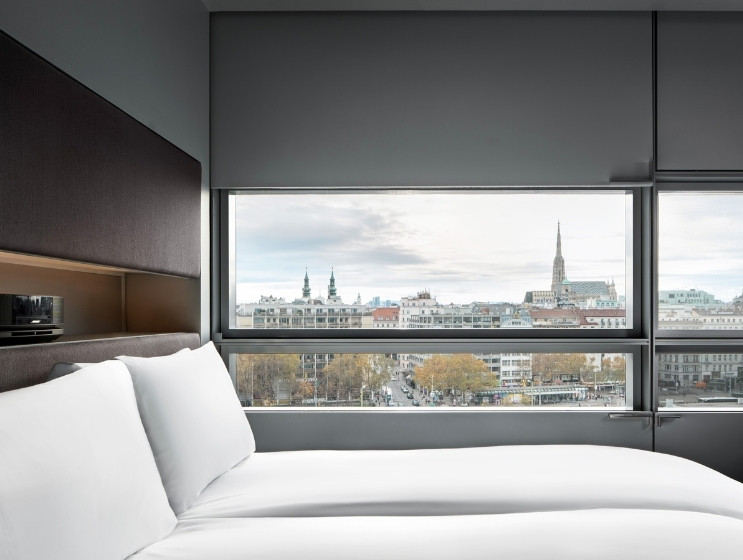 A modern hotel room with a large window showcases a cityscape featuring church spires. The room has a minimalist style with white bedding and a calm atmosphere.
