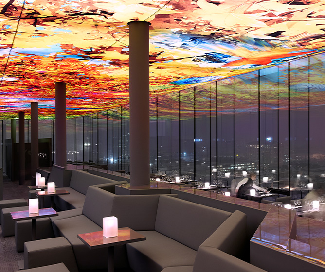 A stunning ceiling art piece illuminates a bar seating area with full length windows displaing the Viennese skyline at night