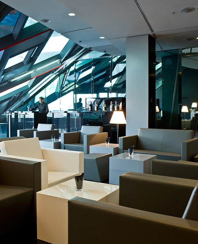 A foyer with square, black and white armchairs. A man sit and the back of the room under a slanted wall. 