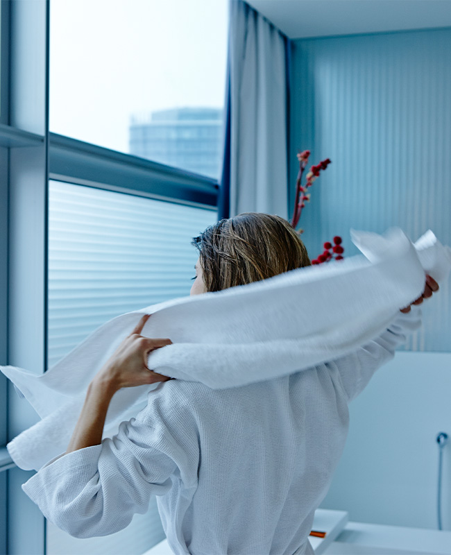 A women in a robe drying her hair with a white towel look outside a large window