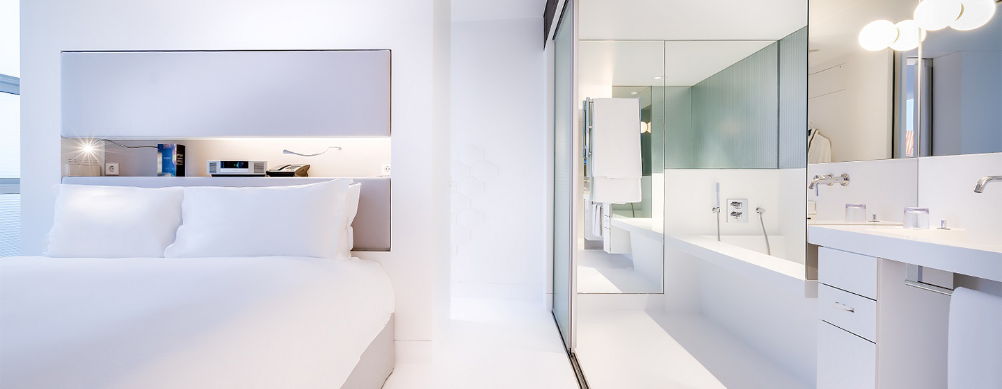 A pristine white hotel room with a double bed and an open sliding showing a white bathroom