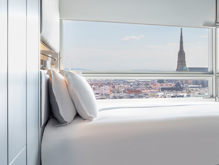 Hotelzimmer mit Blick auf die Stadt, mit einem ordentlich gemachten Bett mit weißer Bettwäsche und zwei weichen Kissen vor einem hellen Fenster.