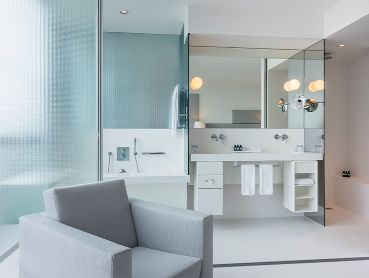 Modernes, minimalistisches Badezimmer mit einem eleganten Waschtisch, einer verglasen Dusche und einer eleganten Beleuchtung, mit einem grauen Sessel im Blickfeld.