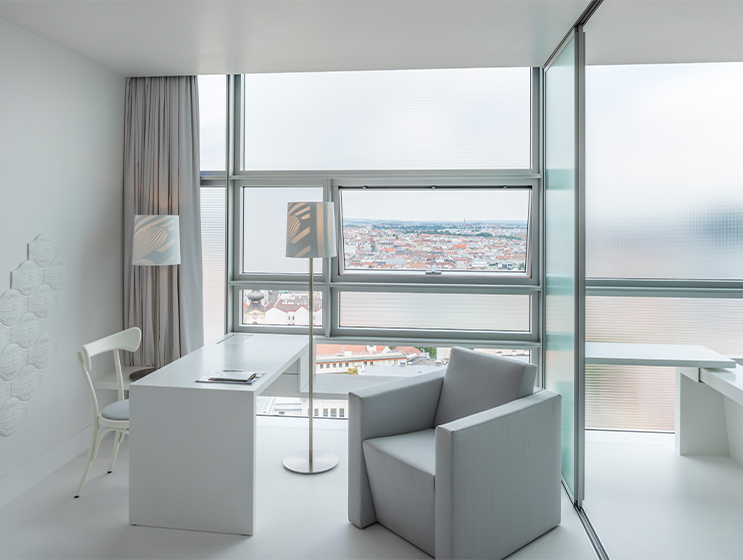 Weißes Hotelzimmer mit großem Fenster, gemütlichem Sessel, kleinem Tisch und sanfter Beleuchtung, das eine ruhige Atmosphäre bietet.