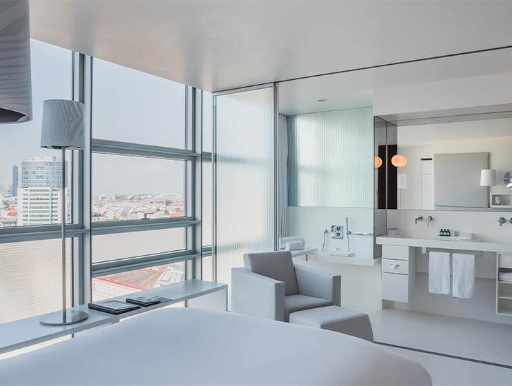 Hotelzimmer mit großen Fenstern, minimalistischer Einrichtung, einer gemütlichen Sitzecke und einem offenen Badezimmer mit eleganten Armaturen.