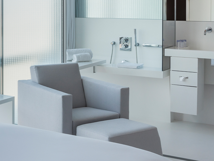 Minimalistisch eingerichtetes Hotelzimmer mit einem hellgrauen Sessel und einer Ottomane, eleganter Einrichtung und heller, luftiger Atmosphäre.