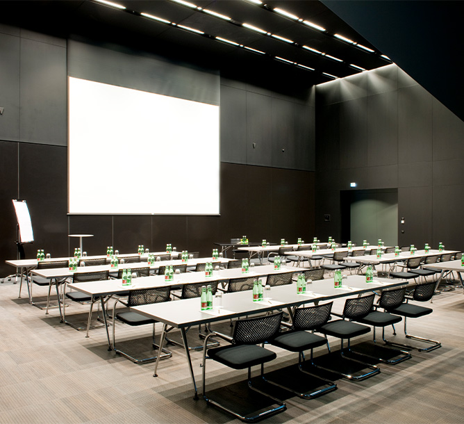 A large, high ceilinged meeting room. Tables and chairs in a classroom set up face large screen hanging on the wall.