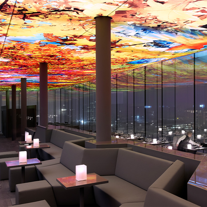 A stunning ceiling art piece illuminates a bar seating area with full length windows displaing the Viennese skyline at night