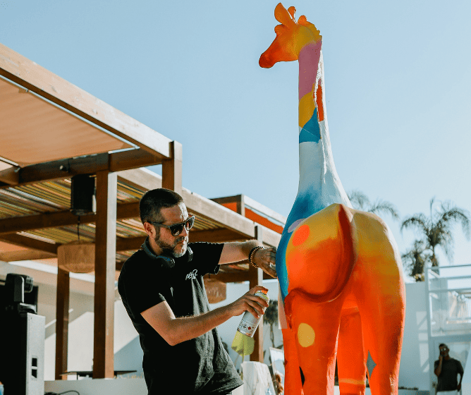 Un artista pinta con spray una escultura de jirafa de colores vivos y multicolores al aire libre, rodeada de palmeras y un entorno moderno.