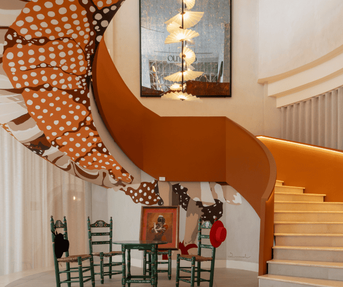 A stylish interior with a spiral staircase, colorful wall art, a green table with chairs, and a framed portrait. Warm lighting enhances the decor.