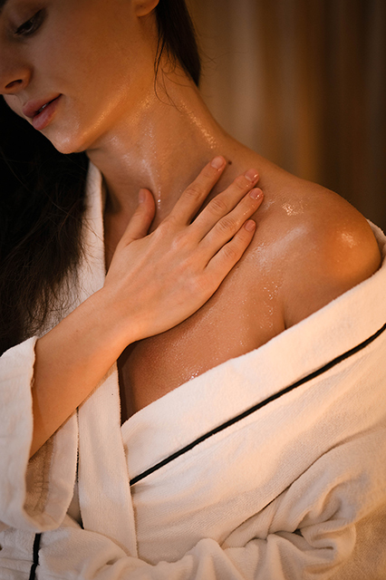 A woman in a white robe gently applies lotion to her neck and shoulder. The scene conveys relaxation and self-care with a warm, soothing light.