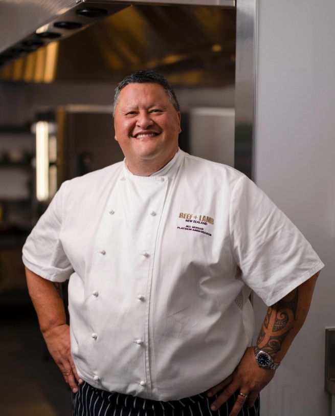 A chef in a white jacket with the "Beef + Lamb New Zealand" logo stands confidently in a professional kitchen setting.