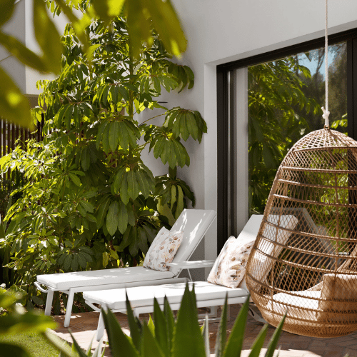 http://A%20patio%20featuring%20a%20swing%20chair%20and%20a%20wicker%20chair,%20surrounded%20by%20greenery%20and%20sunlight.
