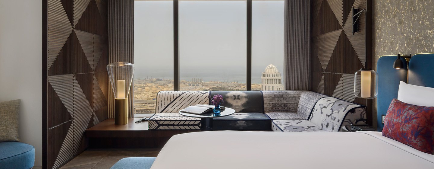 A large bed with blue velvet headrest. A modern sofa bench next to floor-to-ceiling windows looking over the Duba and a reading lap on the side.