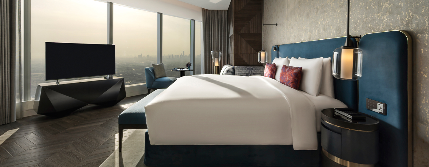 A king size bed in the middle of a hotel room with a blue cushioned bench at the foot of the bed. with a view out onto the Dubai skyline.