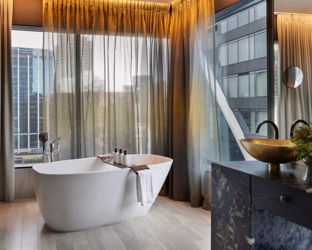 Spacious modern bathroom featuring a freestanding bathtub by large windows, with soft drapes and elegant toiletries displayed.