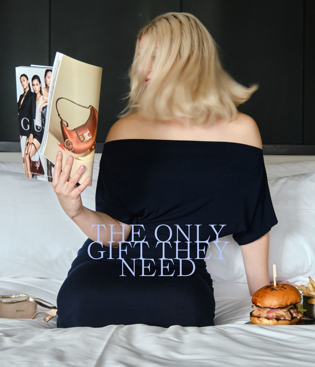 A woman in a black off-shoulder dress sits on a bed, flipping through a magazine featuring handbags. Her hair obscures her face. A burger is beside her. Text reads, “The only gift they need.”