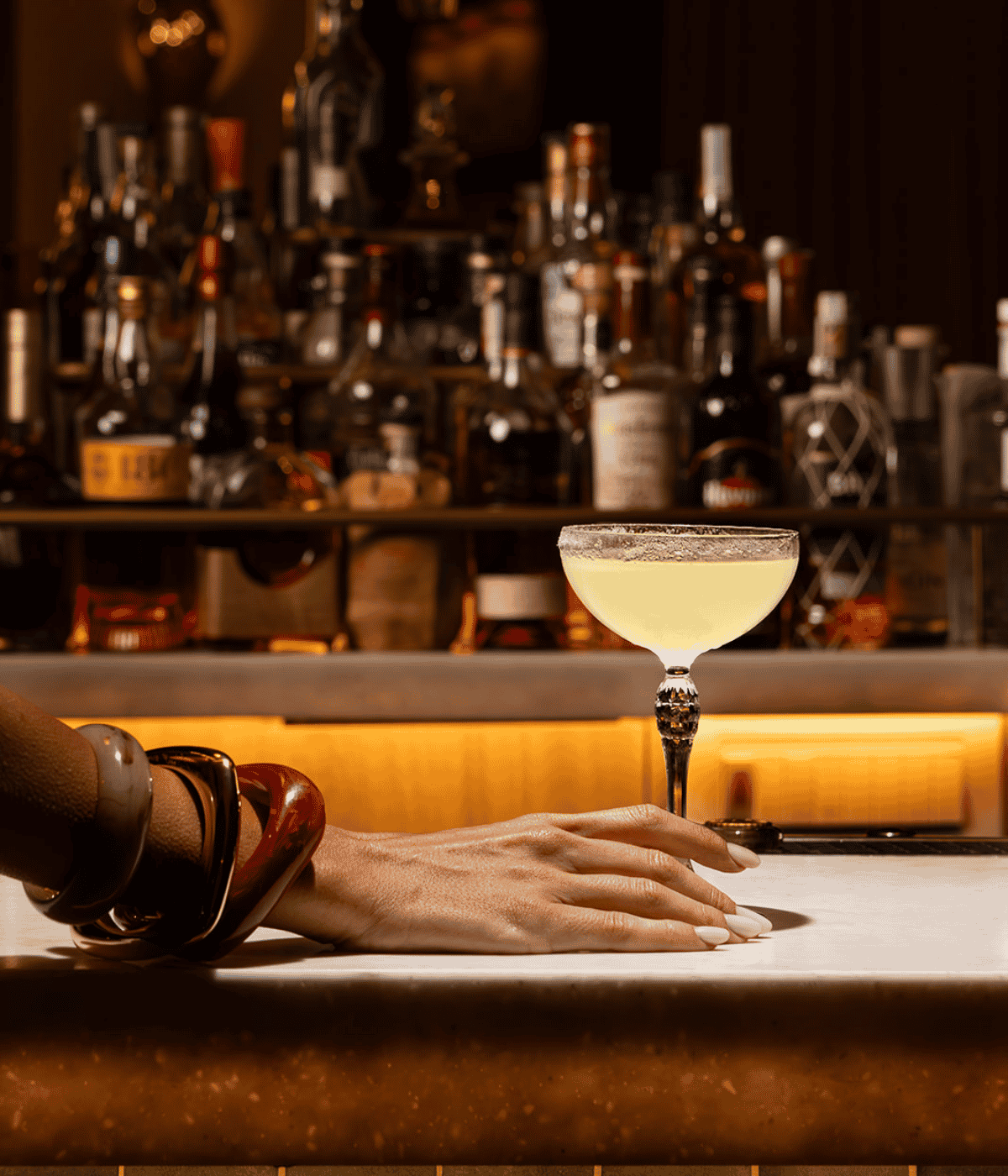 A person holding a cocktail glass, smiling, while seated at a stylish bar with colorful drinks in the background.