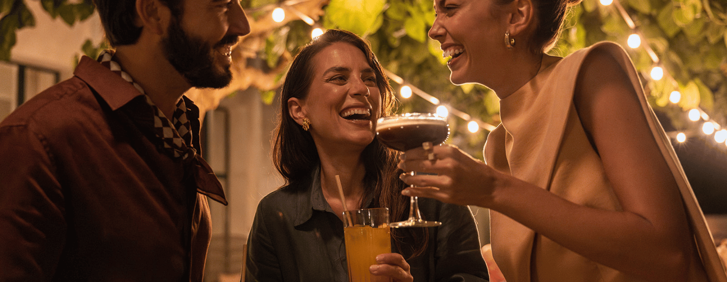 Three friends enjoying drinks and laughing together at an outdoor evening party with string lights and greenery.