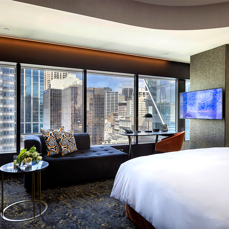 A large open plan hotel room with a bed, desk and blue couch facing a large window overlooking tall buildings