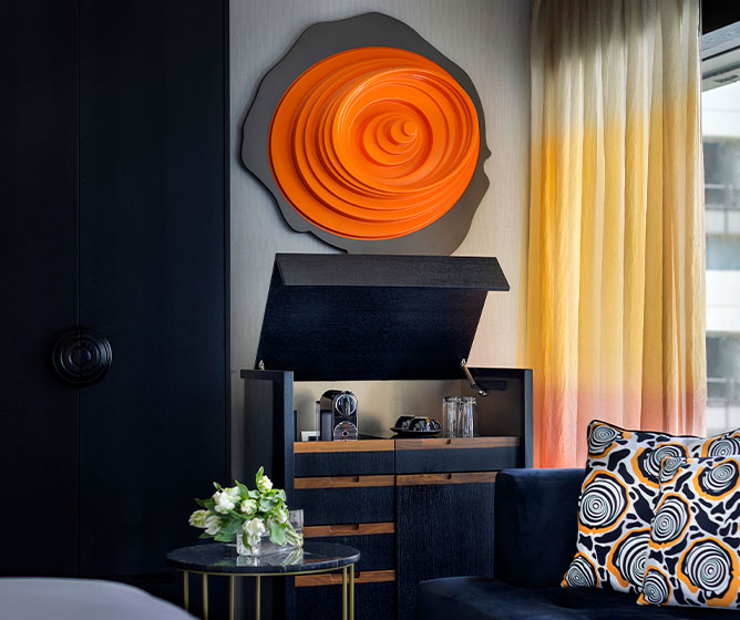 A dark blue couch next to a marbled coffee table and cabinet with a coffee machine and refreshments on it. A large orange emblem on the wall