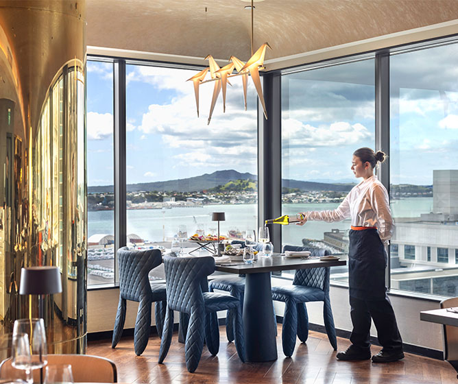 A waitress stands by a restaurant table which overlooks a beautiful of water and mountains.
