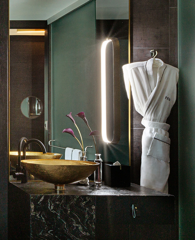 A gold bathroom sink infornt of a large mirror. A white 'his' bathrobe hangs next to the mirror.