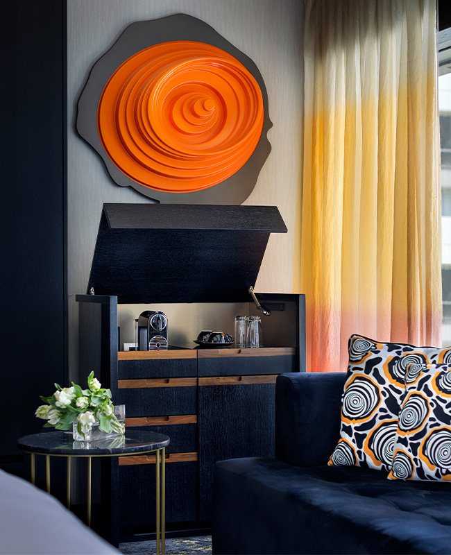 A dark blue couch next to a marbled coffee table and cabinet with a coffee machine and refreshments on it. A large orange emblem on the wall