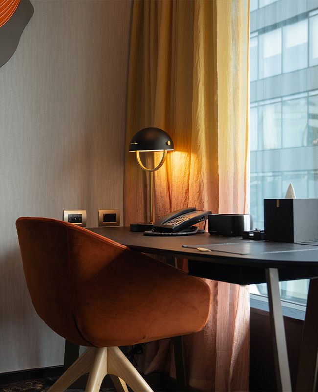 A black desk with desk lamp and telephone and burnt orange desk chair facing a window