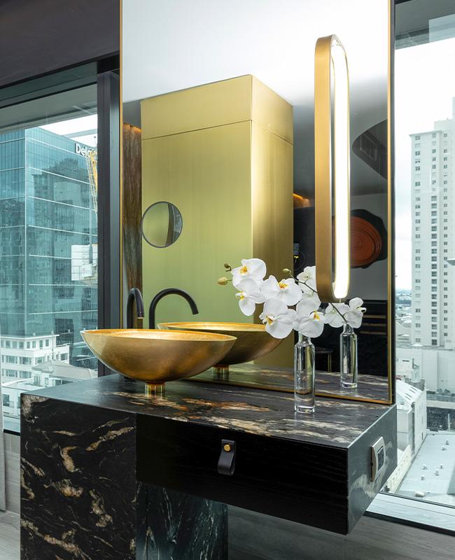 A gold sink tops a marble counter top with a large mirror, behind which is a large window